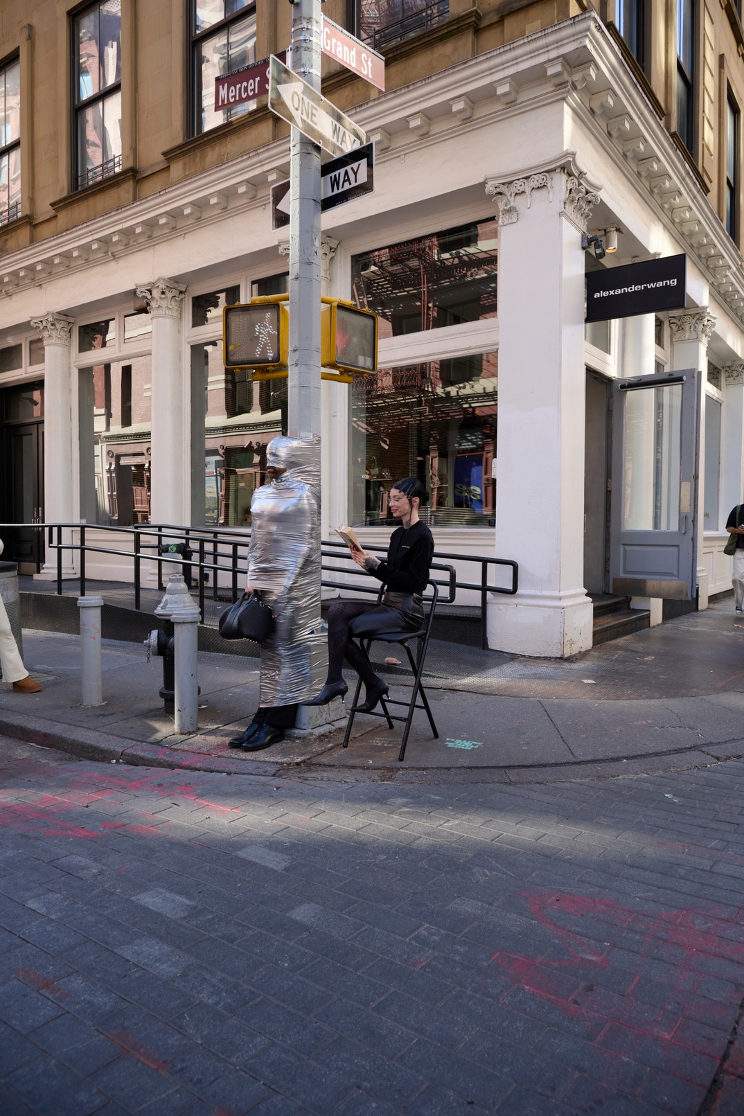 A performer wrapped in silver tape at a SoHo street post holds the Rocco bag, with Sabine Tautou seated beside him outside Alexander Wang.